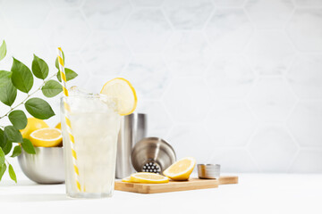 Detox fresh lemon juice cooking - lemonade of ripe lemons with ice in glass with silver bowl, shaker on white table, marble tile wall with green foliage in summer sunlight.