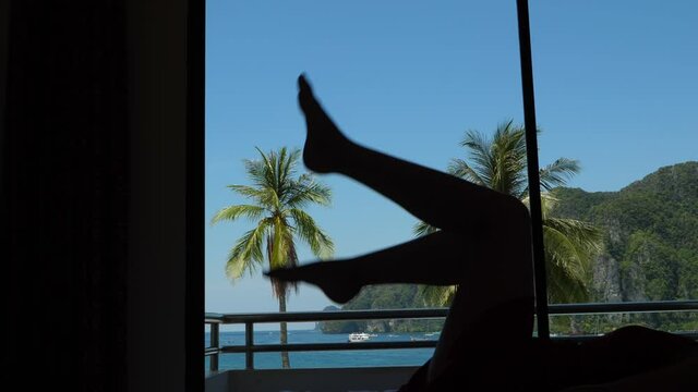 Woman lie on bed, raise up legs and wave feet, enjoy happy holidays time. Silhouetted shot against window, green palm trees and tropical sea bay seen outside. Handheld camera, slow motion shot