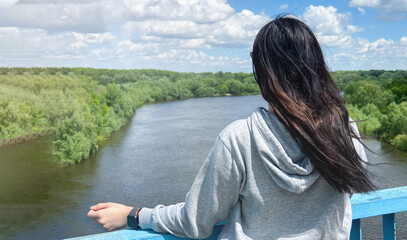 A dark haired caucasian girl stands with her back in a light gray jacket and looks towards the river and the forest