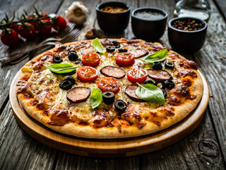 Pizza with white mushrooms, sausage, tomatoes, black olives, parmesan and mozzarella on wooden background
