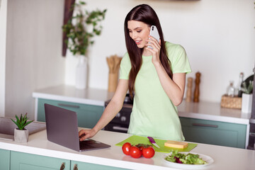 Obraz na płótnie Canvas Photo of adorable busy young woman dressed green t-shirt cooking salad talking typing modern gadgets indoors house home room