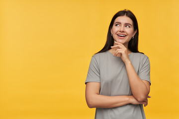 Cheerful attractive brunette young woman in gray tshirt keeps hands folded smiling and looking away to the side at copyspace over yellow background