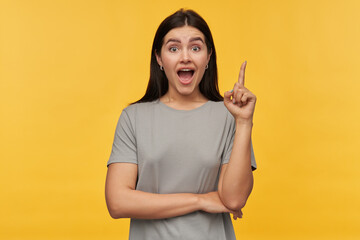 Amazed excited young woman with dark hair and opened mouth in gray tshirt pointing up by finger and...
