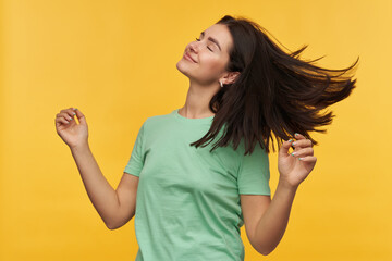 Happy relaxed young woman with dark hair in mint tshirt keeps eyes closed and dancing over yellow background