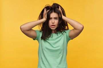 Fototapeta premium Sleepy pretty young woman with dark messy hair in mint tshirt keeps hands on head and feels oversleep isolated over yellow background