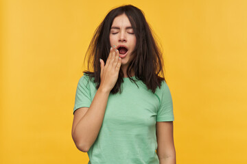 Portrait of tired exhausted brunette young woman in mint tshirt keeps eyes closed and yawning isolated over yellow background