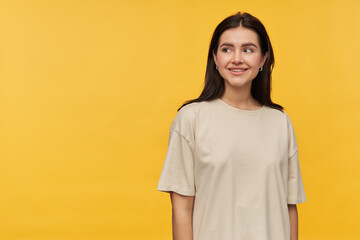 Cheerful beautiful brunette young woman in white tshirt standing and looking away to the side at empty space over yellow background