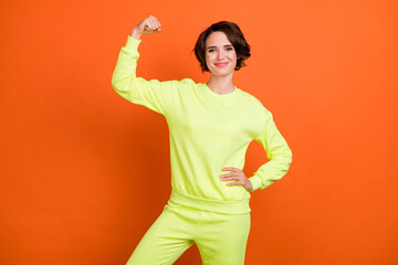 Photo of cheerful young positive happy woman hold hand show muscles flex isolated on orange color background
