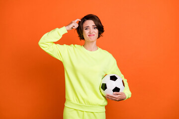 Portrait of attractive puzzled girl holding ball scratching head dont know isolated over bright orange color background