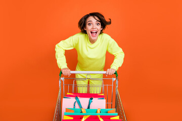 Portrait of attractive amazed cheery girl carrying cart bags things running having fun isolated over bright orange color background