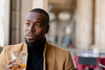 Elegant young black man with serious face looking away holding a glass with juice while sitting in a cafe. City life.