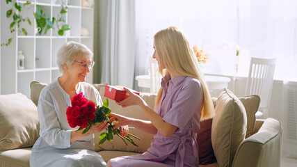Young woman giving a red flowers to his elderly mother in a home interior. Smiling girl with her mother