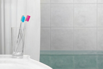 Two toothbrushes in a glass on the sink in the bathroom. Blue and pink toothbrushes. White curtain and white tiles in the bathroom. Space for text.