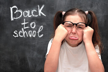 A schoolgirl with ponytails grimaces against the background of a black chalkboard with the words "Back to School" written on it. The girl is unhappy that she needs to go to school again.