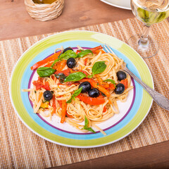 Portion of vegetarian pasta, spaghetti with bell pepper and olives with basil leaves on the table with a glass of white wine