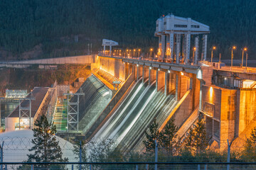 Illuminated hydroelectric power plant behind barbed wire. Water discharge. Picture on long...