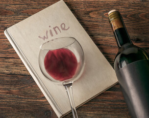 A glass of red wine lies on a book with the inscription "wine". And next to it, on a wooden textured table, lies a bottle.