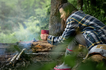 Young woman resting near the fire with a cup of warming drink in the forest.
