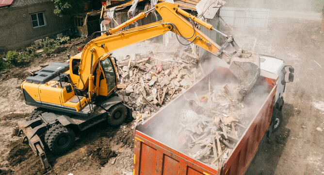 Excavator breaks building and loads construction waste into truck with its bucket. Demolition of building.