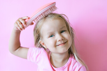 little girl combing her hair. Beauty and childhood concept. Girl on a pink isolated background combing her hair with a pink comb - obrazy, fototapety, plakaty