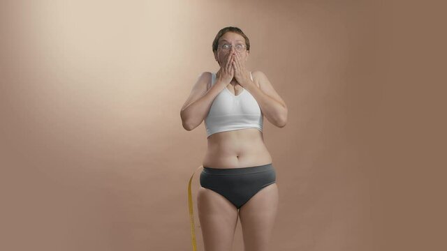 Full figured girl in white bra and black briefs with measure tape to know the volume of her waist. Surprise face expression. Beige background slow motion high quality video.