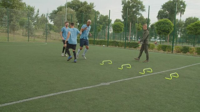 Tree soccer players in football uniform alternately performing jump barriers exercise outdoors at daybreak, while handsome young trainer controlling, urging team to move faster looking at hand watch