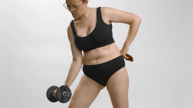 Young beautiful caucasian plus size model in sports underwear and glasses with short hair training with weight for strength and taking a bite of cookie. White background slow-motion studio video.