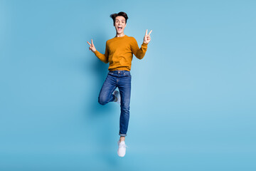 Full length photo of cool brunet young guy jump show v-sign wear sweater jeans isolated on blue color background