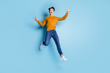 Obraz premium Full size photo of crazy brunet millennial guy jump show v-sign wear sweater jeans isolated on blue color background