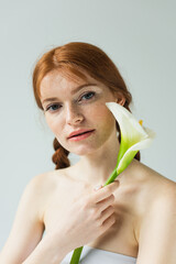 Freckled woman holding calla lily and looking at camera isolated on grey