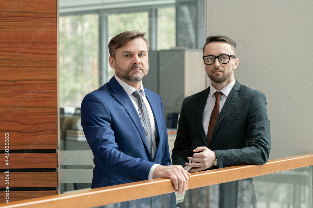 Poster portrait of two confident businessmen in elegant suits looking at camera while standing in the corri