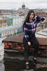 An Armenian girl with flowing long black hair in a checkered shirt sits on a rooftop in the center of St. Petersburg