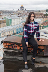 An Armenian girl with flowing long black hair in a checkered shirt sits on a rooftop in the center of St. Petersburg