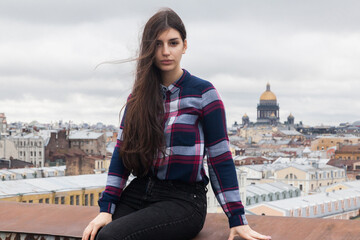 An Armenian girl with flowing long black hair in a checkered shirt sits on a rooftop in the center of St. Petersburg