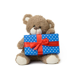 cute brown teddy bear holding a box wrapped in blue paper and red silk ribbon on white isolated...