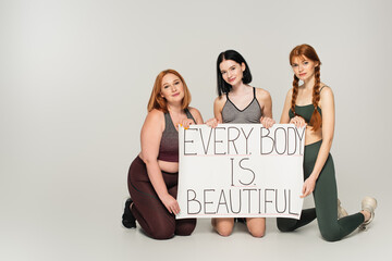 Body positive women in sportswear holding placard with every body is beautiful lettering on grey background