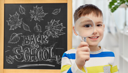 education, learning and people concept - happy little student boy looking through magnifying glass over chalkboard with back to school lettering on background