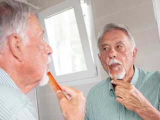 Elderly Caucasian man takes care of his personal hygiene in front of the mirror in the bathroom