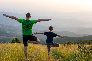 Father and son doing yoga Tree pose at top of mountain at sunset. Happy little boy playing with his father practicing child yoga pose. Family are exercising or yoga with mountain view.