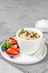 Mug cake with chocolate chips, fresh berries and cream on a gray concrete background. Copy space.