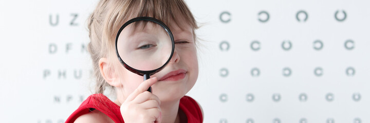 Little girl with magnifying glass standing on background of table for eye test