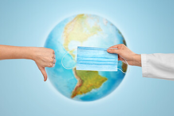 medicine, health protection and healthcare concept - hand of doctor with medical mask and patient showing thumbs down over earth globe on blue background