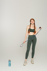 Cheerful sportswoman with freckles holding jump rope near sports bottle on grey background