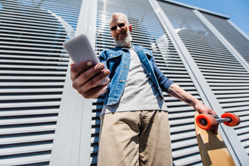 low angle view of pleased middle aged man in sunglasses using smartphone and holding longboard