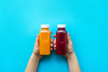 Female hands holding plastic bottles with orange juice or red smoothie on blue background. The...