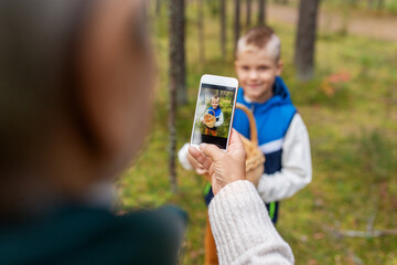 picking season, leisure and people concept - grandmother with smartphone photographing happy smiling grandson with mushrooms in basket in forest