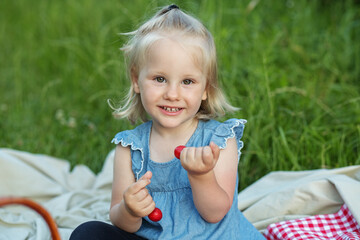 A cheerful girl with blonde hair is eating cherries in nature, trying berries on a picnic