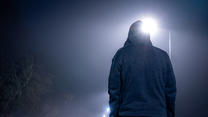 Looking up from behind at a mysterious, moody, hooded figure. On a foggy winters night, with street lights behind