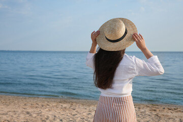 Beautiful young woman wearing straw hat on beach, back view. Stylish headdress