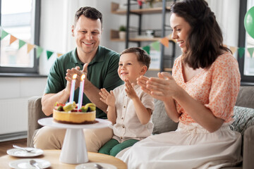 family, holidays and people concept - portrait of happy mother, father and little son with four candles on birthday cake sitting on sofa at home party